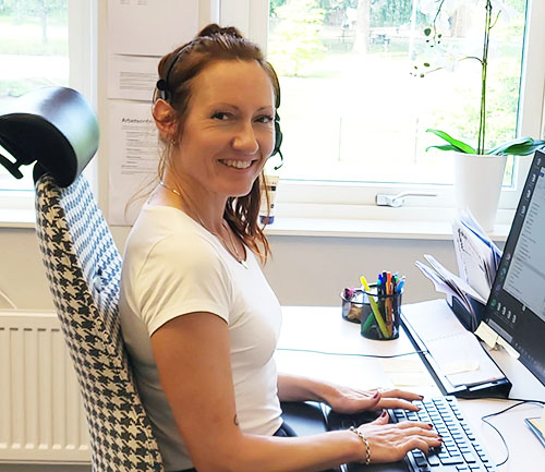 En kvinna sitter leende vid ett skrivbord och jobbar på datorn. 